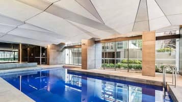 A modern indoor swimming pool with clear glass walls, a tiled ceiling, and a metal ladder.