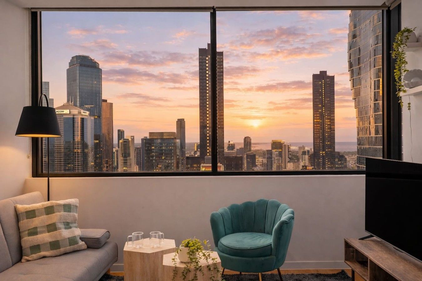 A room with a large window overlooking a city skyline at sunset, a teal armchair, a small side table, and a sofa with a checkered throw pillow.
