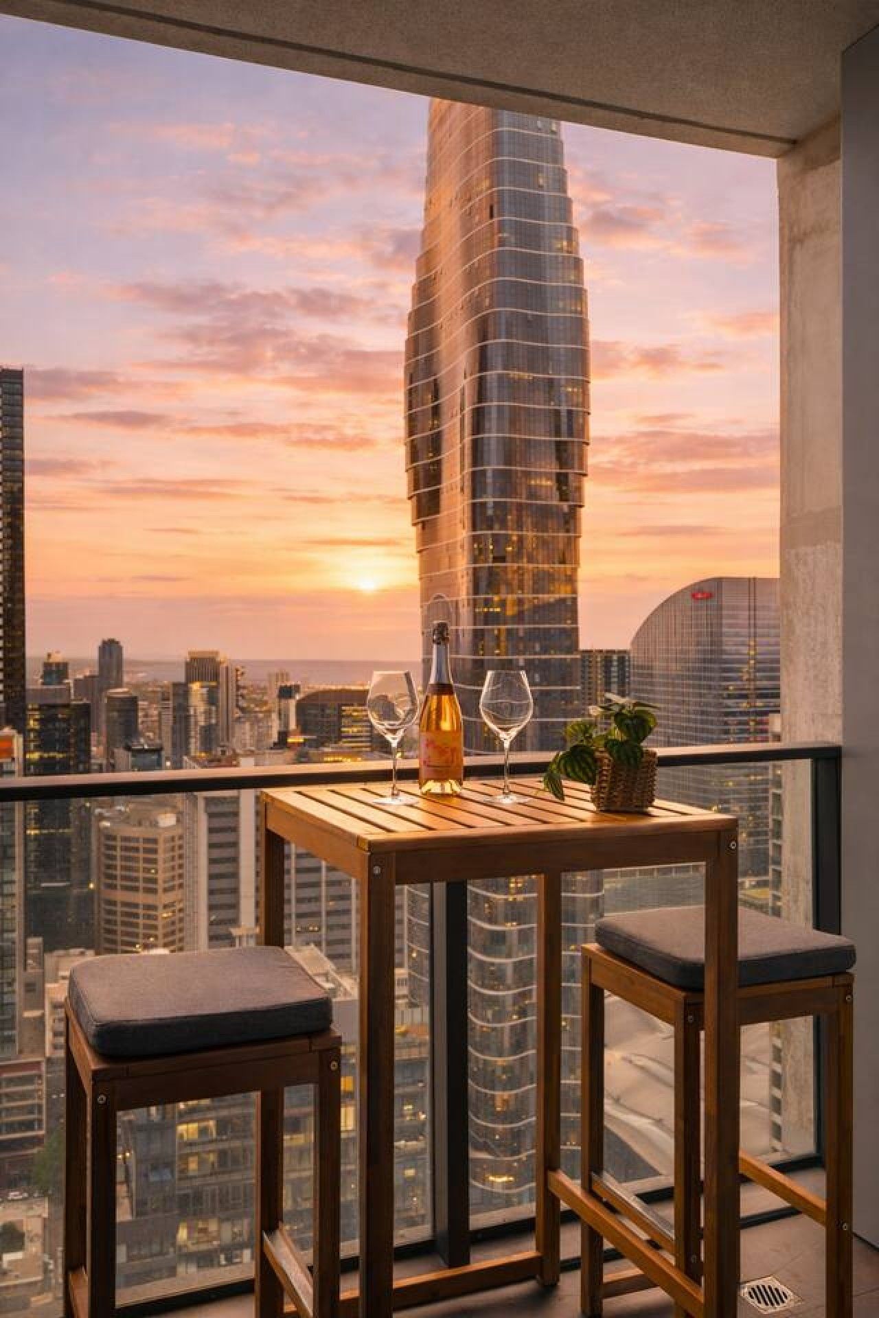 Un balcón con una mesa, dos sillas, una botella de vino y una copa, con vistas a un paisaje urbano que incluye un edificio alto al fondo.