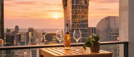 Un balcón con una mesa, dos sillas, una botella de vino y una copa, con vistas a un paisaje urbano que incluye un edificio alto al fondo.