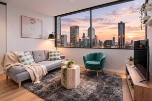 A modern living room with a grey sofa, teal armchair, and a city view. - Golden Hour · 2BR · Level 42 Skyline + Pool (Melbourne)