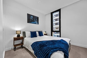 A bedroom with a bed, a nightstand, a lamp, a window, and a painting on the wall. - Designer 2BR · Heart of CBD · Pool + Free Trams (Melbourne)