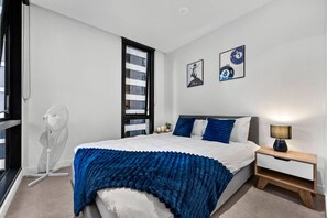 A bedroom with a bed, a bedside table with a lamp, a window, and a wall-mounted picture. - Designer 2BR · Heart of CBD · Pool + Free Trams (Melbourne)