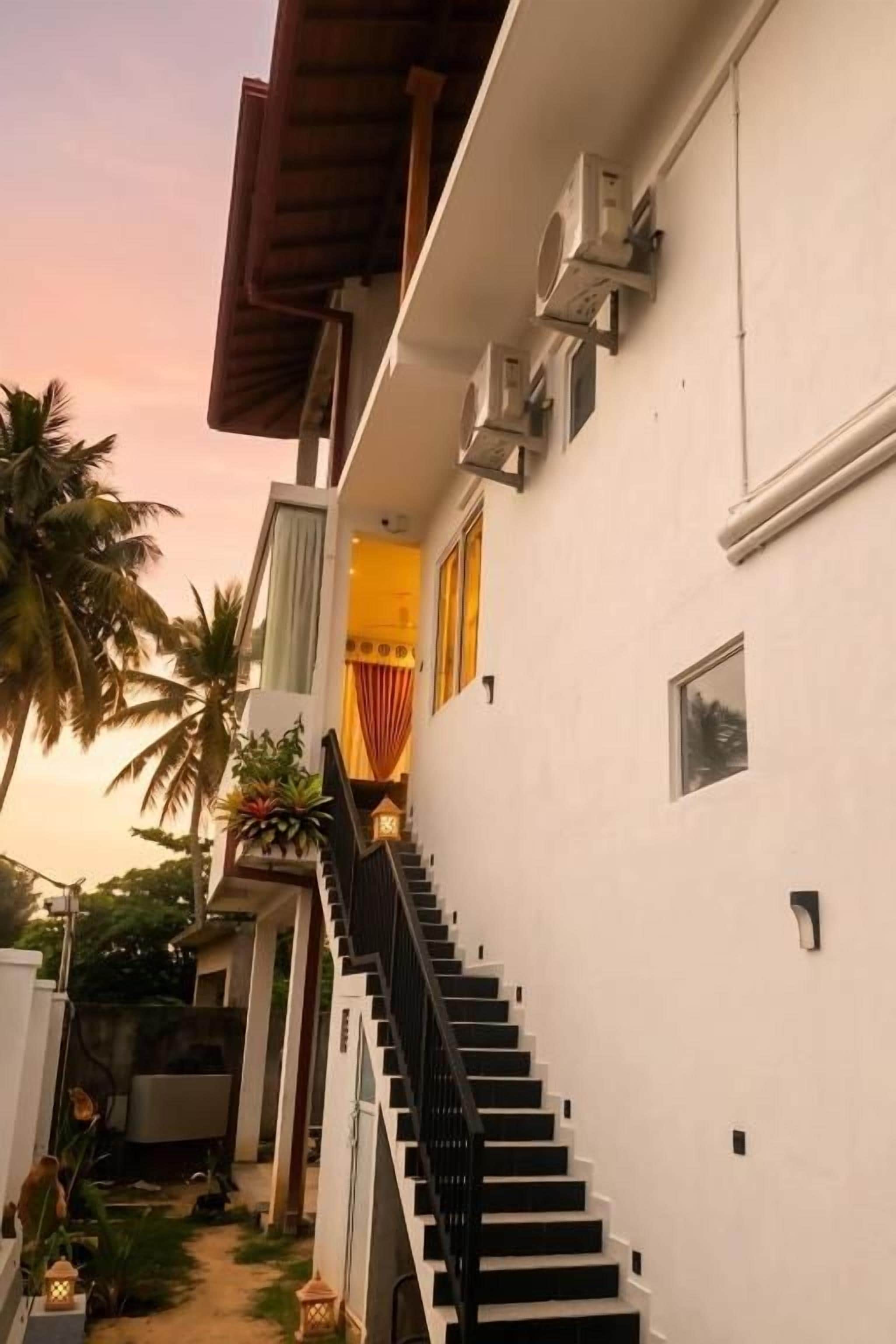 A white building with a black staircase leading to a door, surrounded by palm trees.
