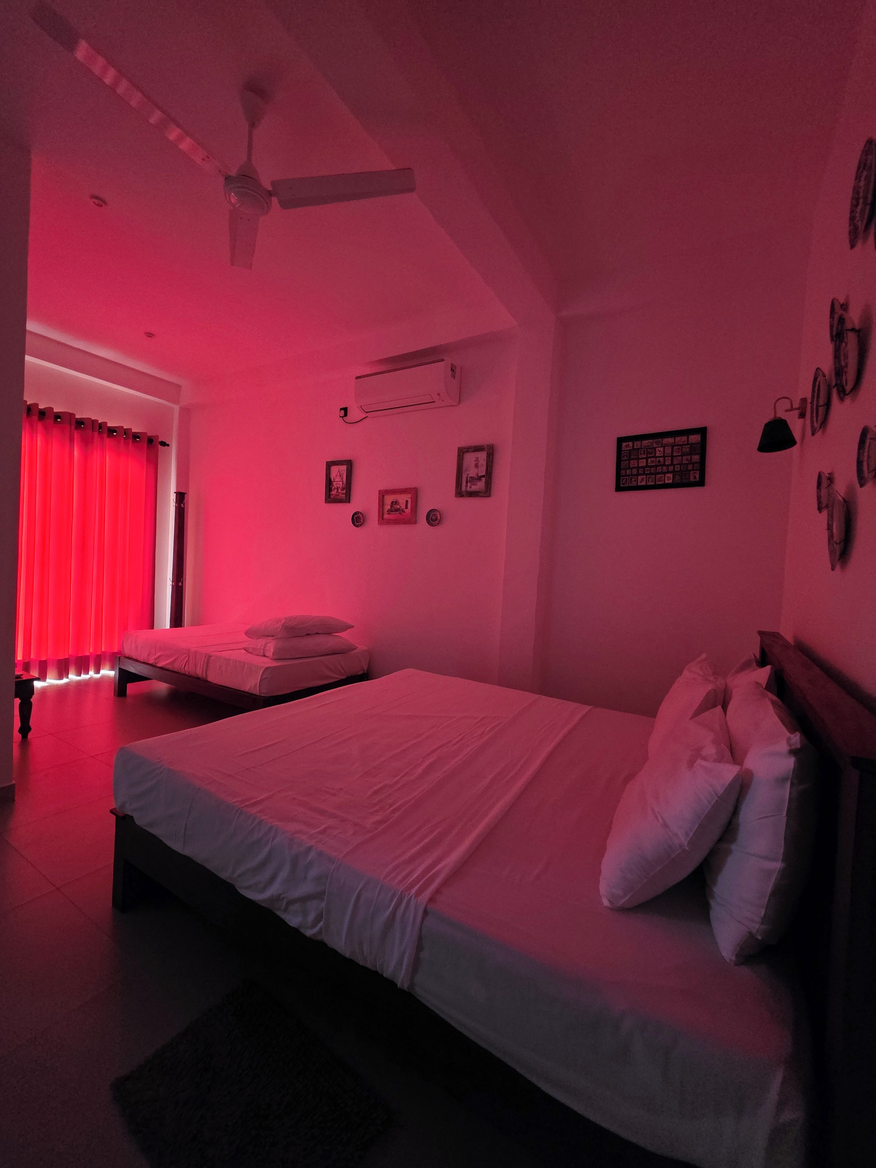 A bedroom with a bed, a ceiling fan, an air conditioning unit, and a window with red curtains.