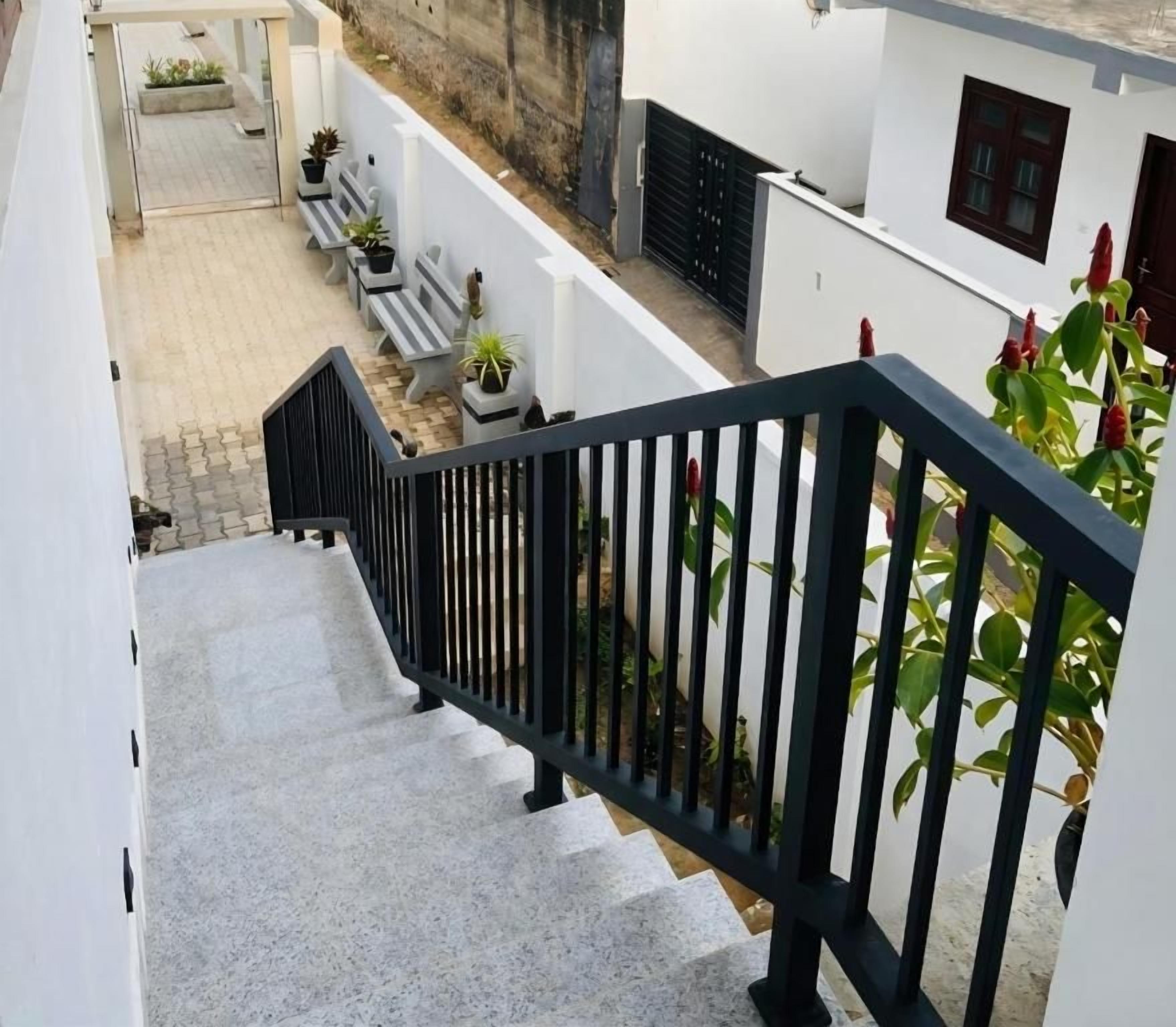 Un escalier avec une rampe noire menant à un espace extérieur abrité, aménagé avec des bancs et des plantes en pot.