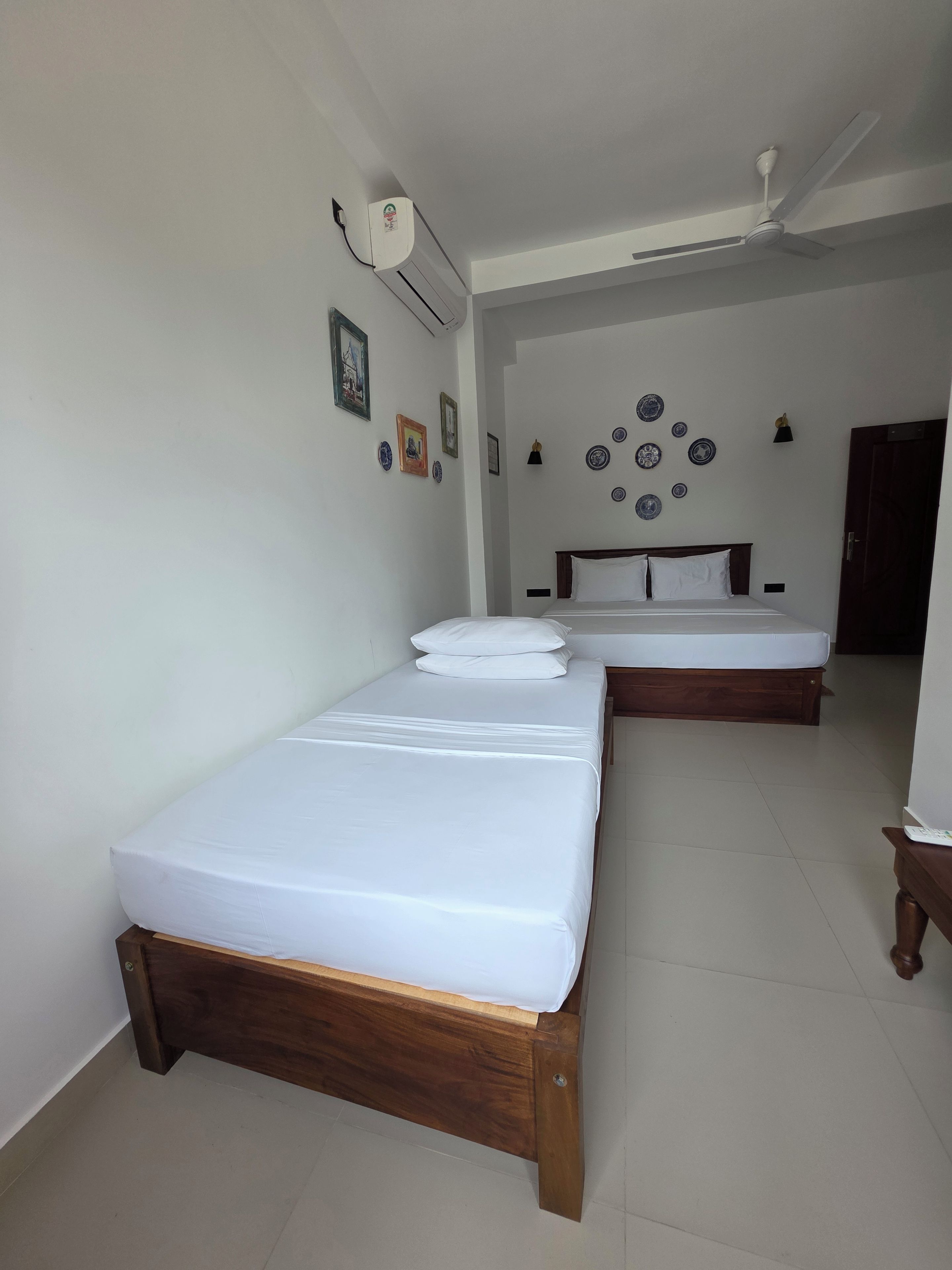 A hotel room with two single beds, a ceiling fan, and a wall-mounted air conditioner.