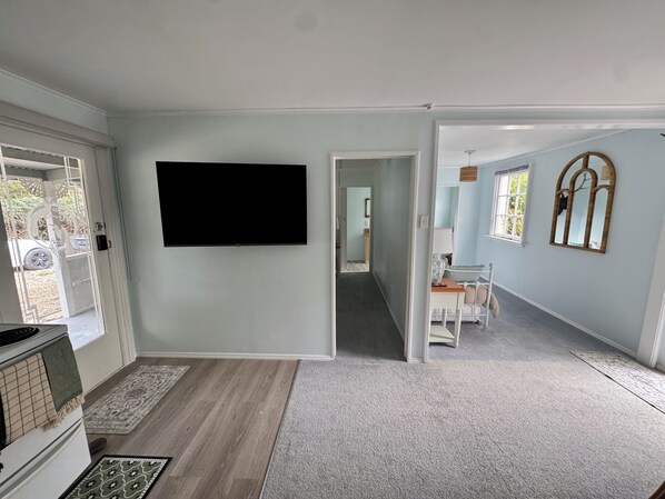 A room with a flat-screen TV mounted on the wall, a kitchen area with a stove, and a hallway leading to another room.