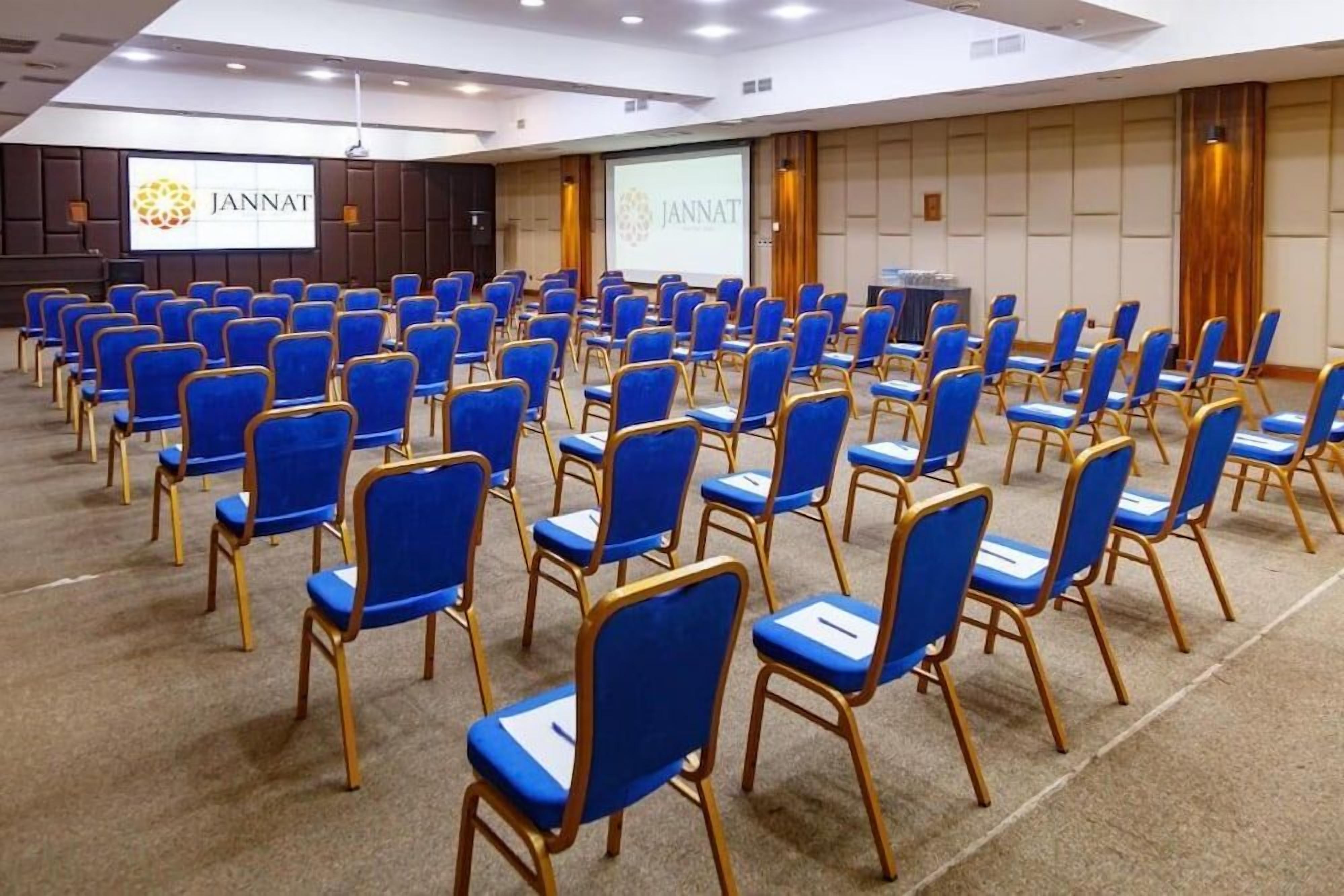 A conference room with rows of blue chairs, a presentation screen, and a JANNAT logo.