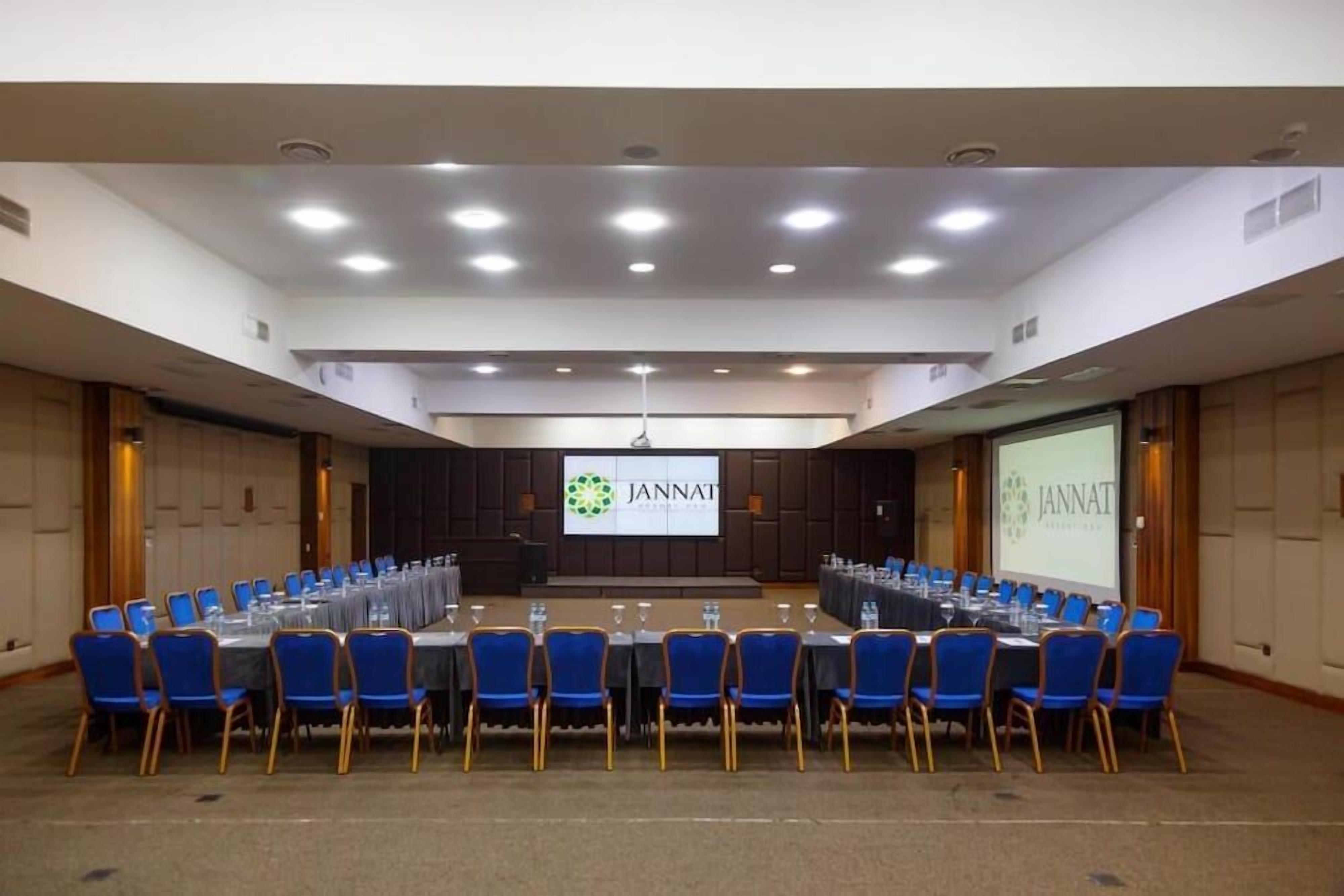 Sala de conferência com uma mesa comprida, cadeiras azuis e tela com a marca JANNAT.