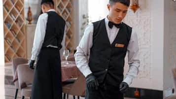 A waiter in a black vest and bow tie standing at a table with a white cup and saucer.