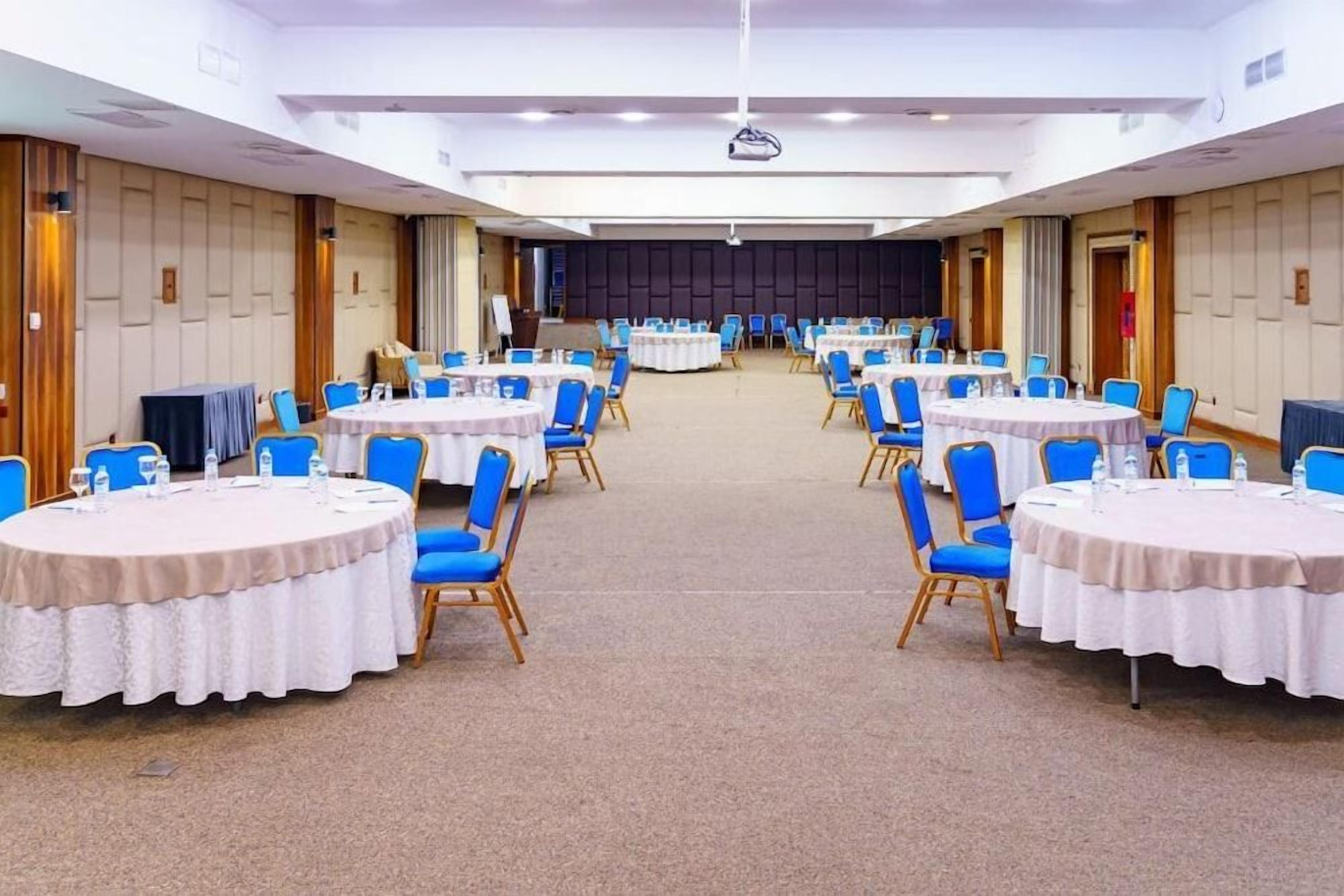 Un salón de conferencias con varias mesas redondas, sillas azules y un escenario al frente.