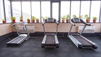 A gym with three treadmills aligned in a row, large windows with potted plants, and a visible air conditioning unit.