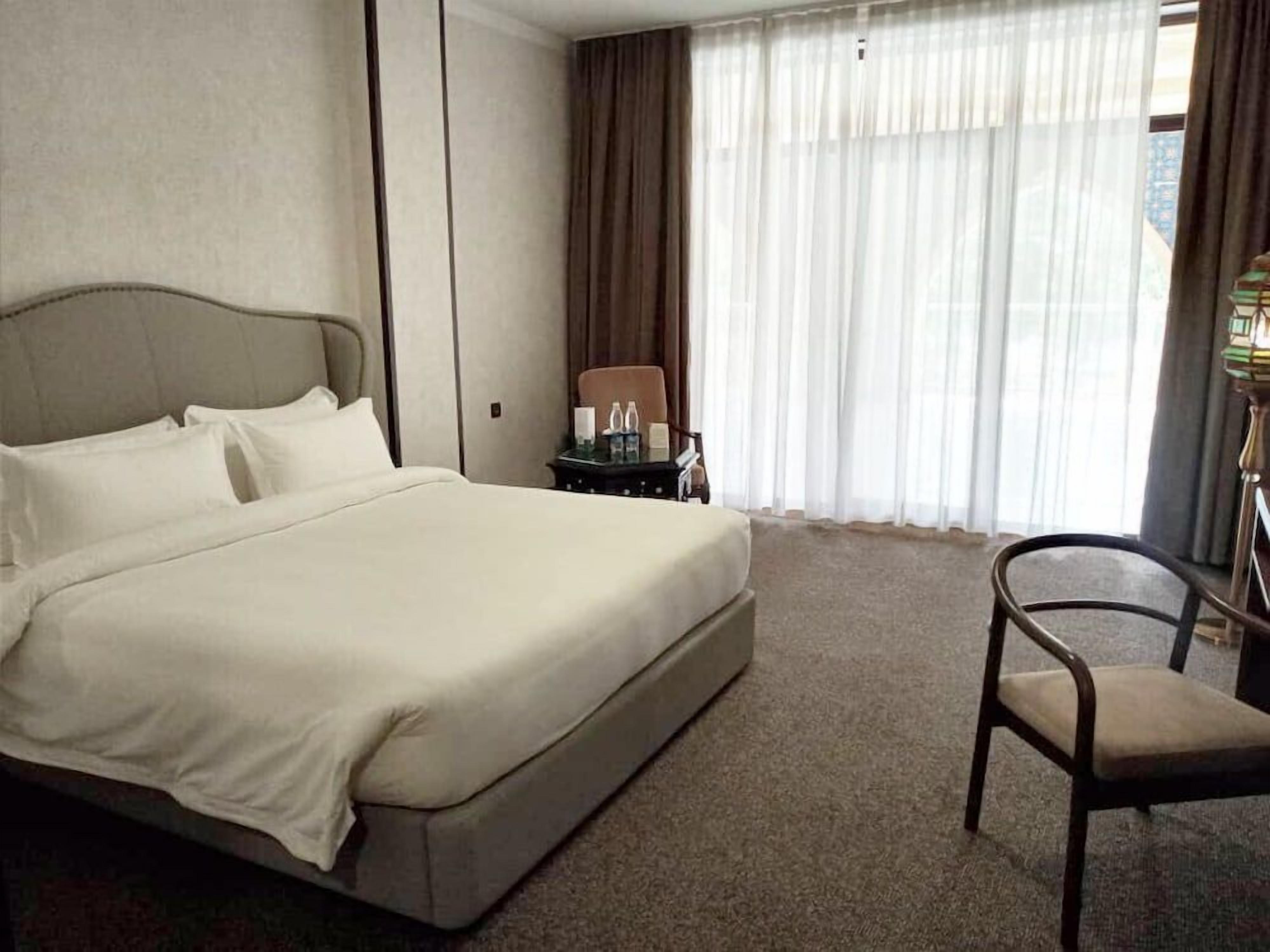 Habitación de hotel con una cama grande, una silla, una mesita con lámpara y vistas a través de cortinas translúcidas.