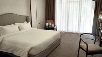 Habitación de hotel con una cama grande, una silla, una mesita con lámpara y vistas a través de cortinas translúcidas.