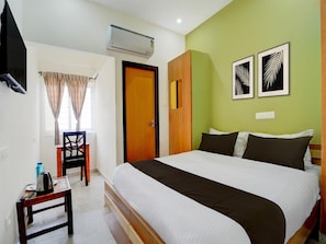 A hotel room with a bed, a desk, a chair, an air conditioning unit, and two framed pictures on the wall.