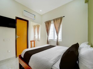 A hotel room with a bed, a TV mounted on the wall, an air conditioning unit, a window with curtains, and a door.