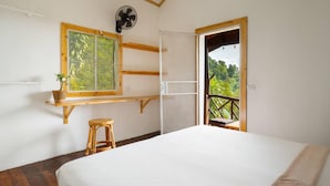 Een slaapkamer met een bed, een raam met uitzicht, een ventilator, een kruk en een deur naar een buitenbalkon.
