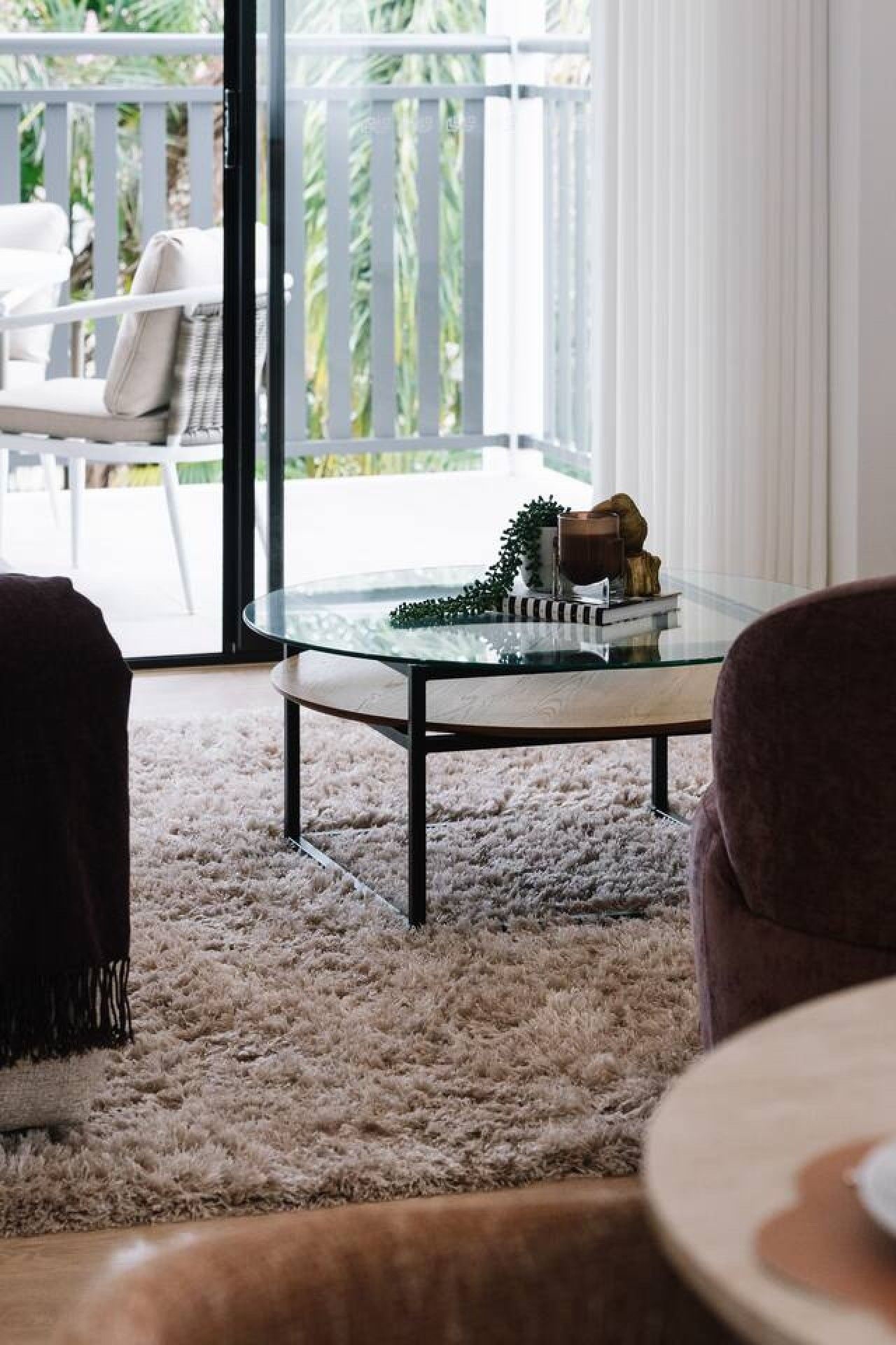 A living room with a glass coffee table, a white shag rug, and a view of a balcony with outdoor furniture.