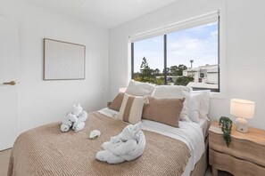 Un dormitorio con una cama grande, una mesita de noche con lámpara y una ventana con vistas a edificios y árboles.