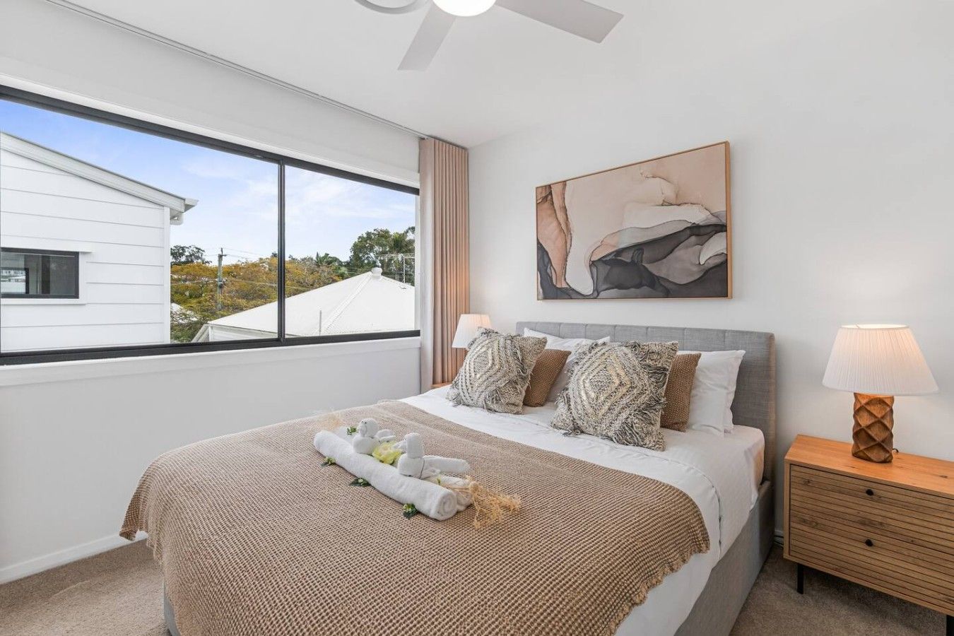 A bedroom with a bed, a nightstand, a lamp, a window with a view, and a painting on the wall.