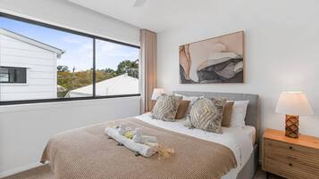 A bedroom with a bed, a nightstand, a lamp, a window with a view, and a painting on the wall.