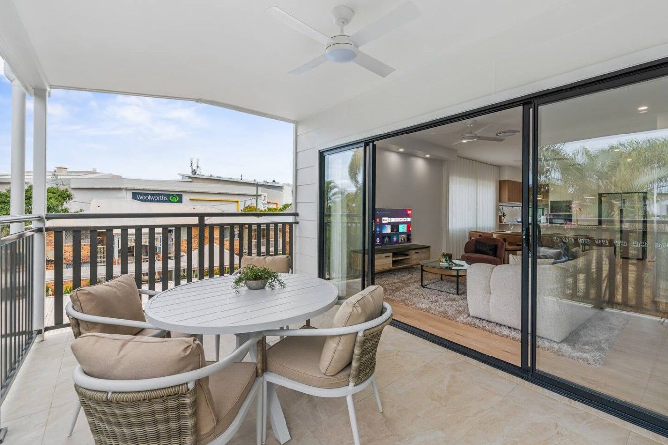 A balcony with wicker chairs and a table, a sliding glass door leading to a living area with a TV, and a ceiling fan.