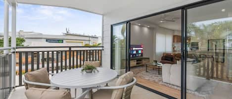 A balcony with wicker chairs and a table, a sliding glass door leading to a living area with a TV, and a ceiling fan.