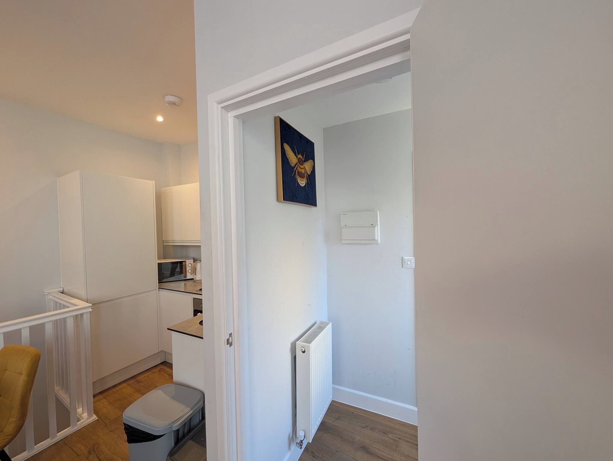 A compact kitchen with white cabinetry, a microwave, and a kettle. There's a radiator on the wall and a trash bin in the corner.