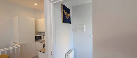 A compact kitchen with white cabinetry, a microwave, and a kettle. There's a radiator on the wall and a trash bin in the corner.