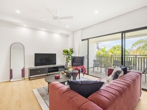 A modern living room with a large flat-screen TV, a sofa, a coffee table, and a balcony with outdoor furniture. - Brand-New Smart Home |3 Min Walk to Train & Shops (Oxley)