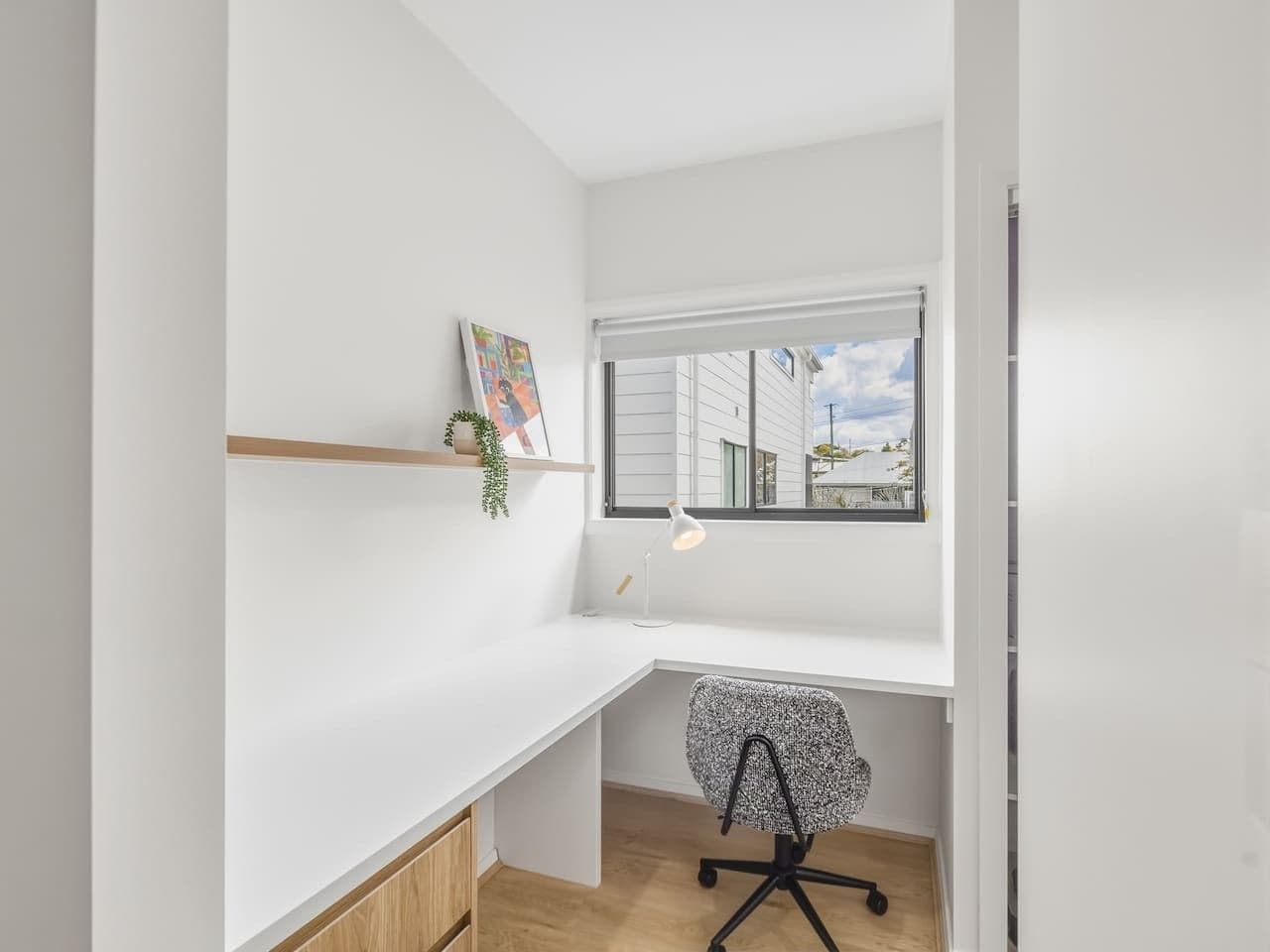 A small, bright room with a white desk, a chair, a window with a view of buildings, and a shelf with a plant.