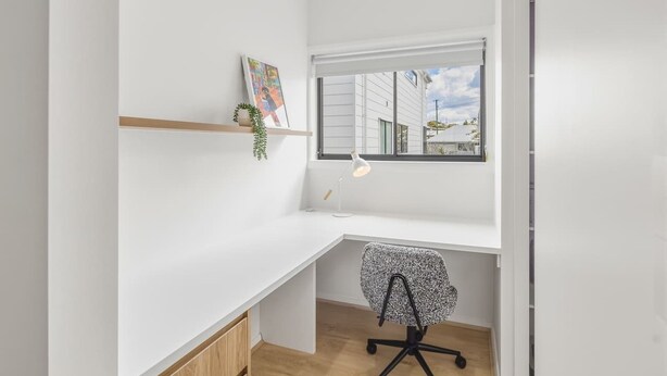 Una habitación pequeña y luminosa con un escritorio blanco, una silla, un ventanal con vistas a edificios y una estantería con una planta.