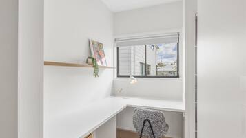 A small, bright room with a white desk, a chair, a window with a view of buildings, and a shelf with a plant.