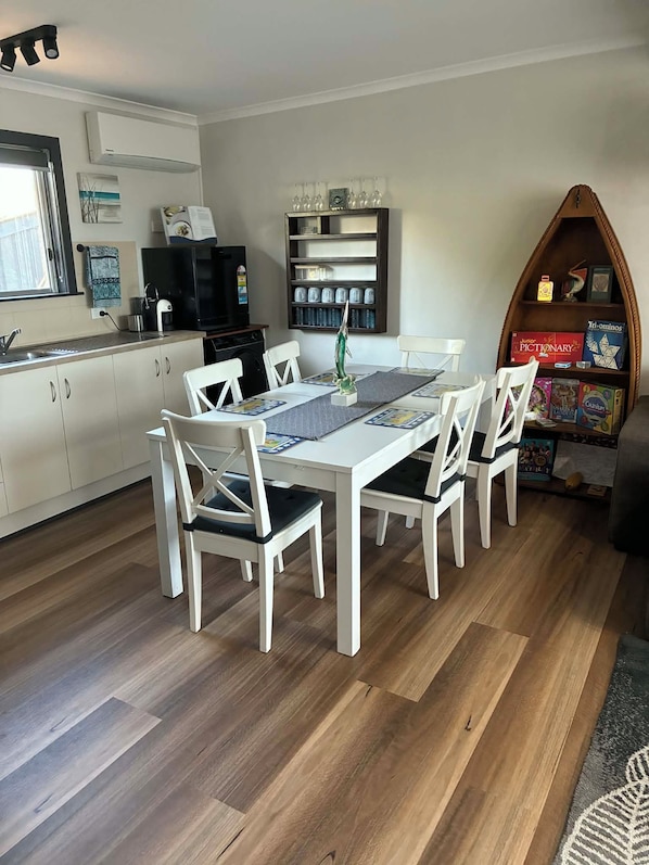 A dining area with a white table and chairs, a kitchen in the background, and a bookshelf with board games. - 2-bedroom studio in the heart of Kingscote (KINGSCOTE)