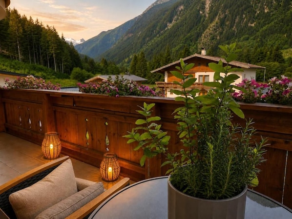 A wooden deck with a sofa and two table lamps, overlooking a mountainous landscape with a house and blooming flowers. - Alpenchalet Vital (Neustift im Stubaital)