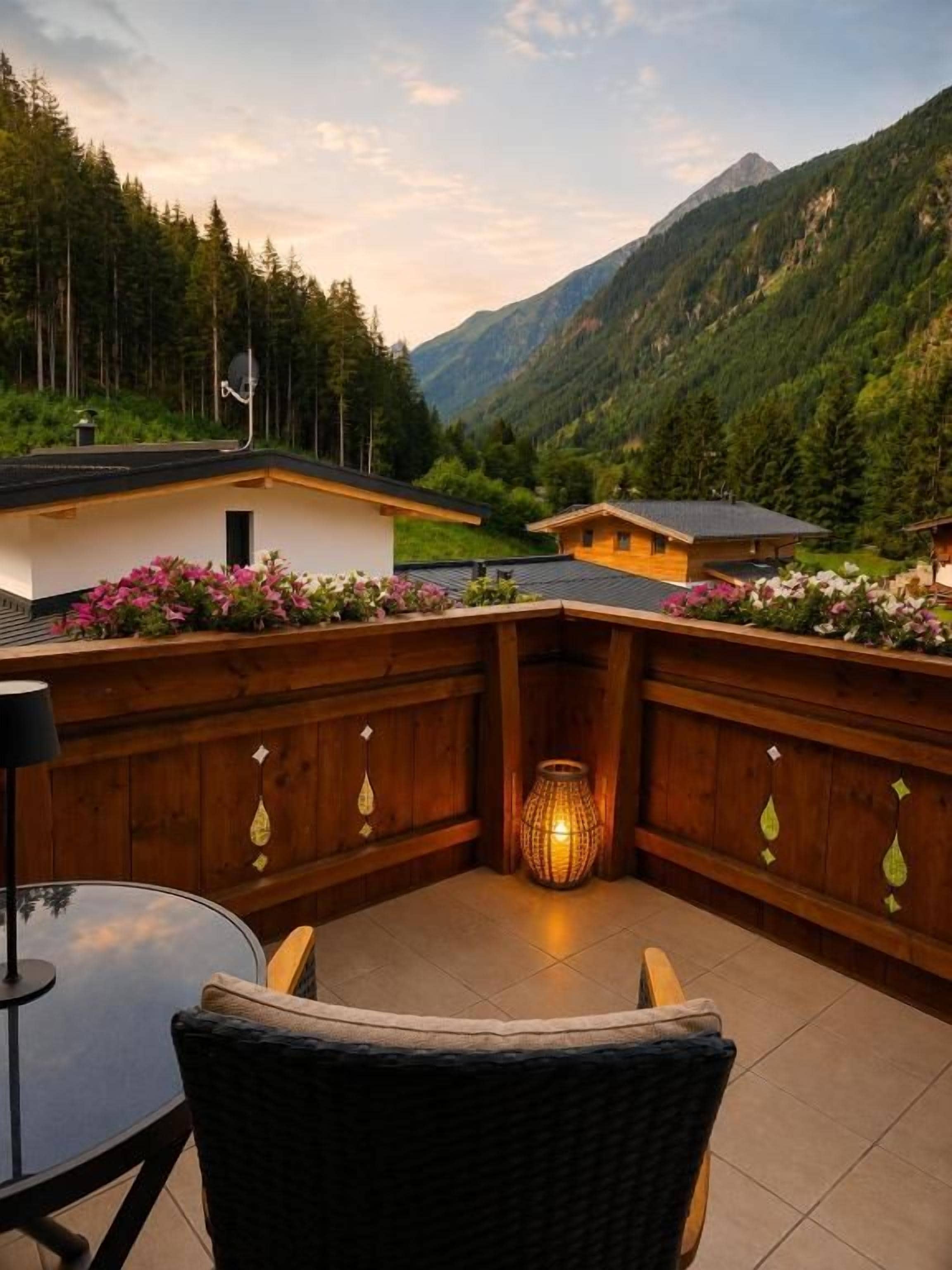 Une terrasse en bois avec une chaise en osier et une table, d’où l’on peut admirer un paysage montagneux avec un petit bâtiment et une lampe.