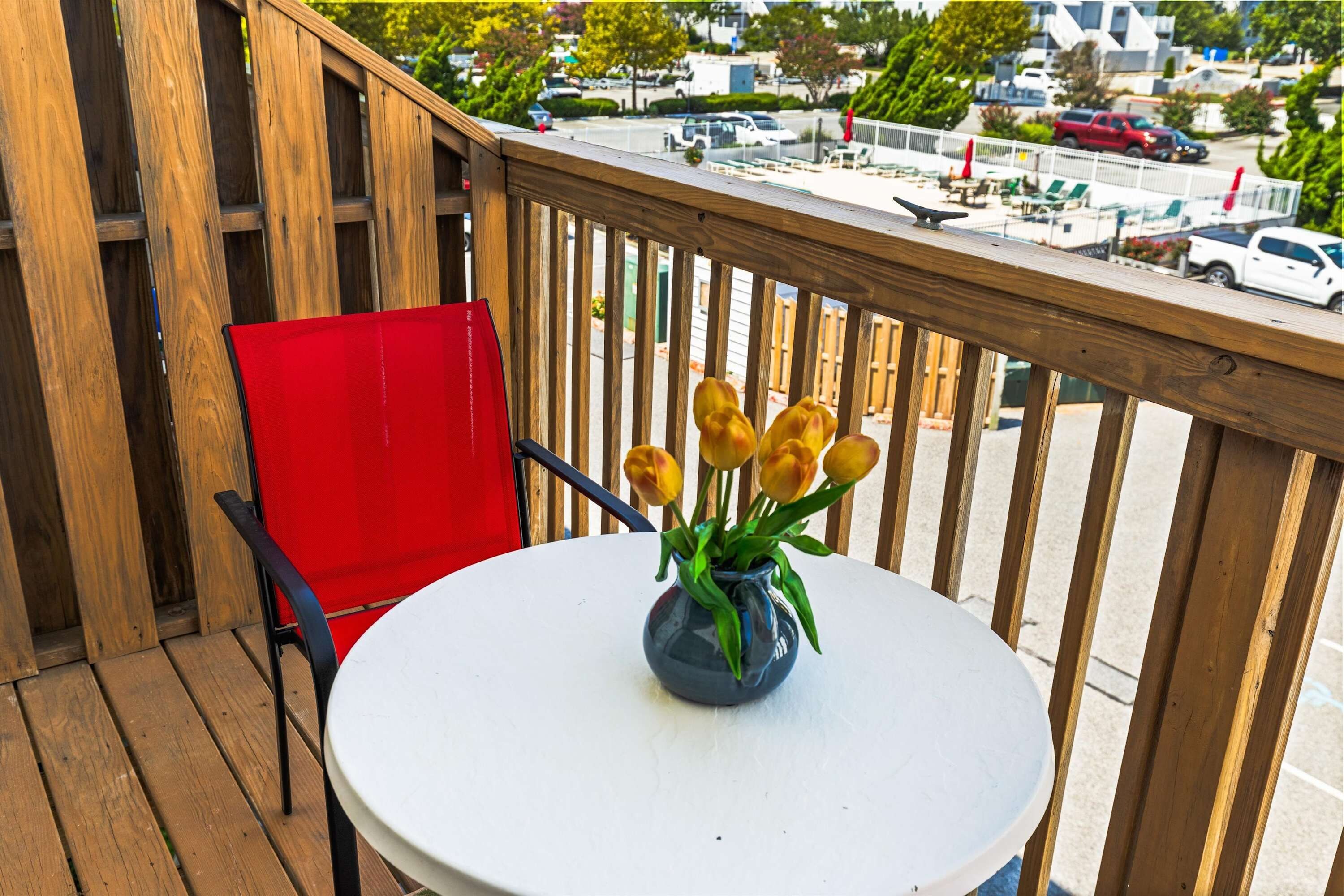 A wooden deck with a round table and a red chair, a vase with yellow flowers, and a view of a parking lot and buildings in the background.