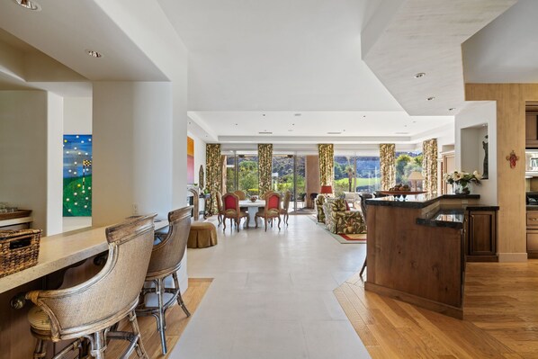 A spacious dining area with a long table and chairs, a kitchen area with a wooden counter, and large windows offering a view of the outdoors. - The Reserve - Luxury 4-Bedroom Retreat Plus Office in Peaceful Indian Wells (Indian Wells)