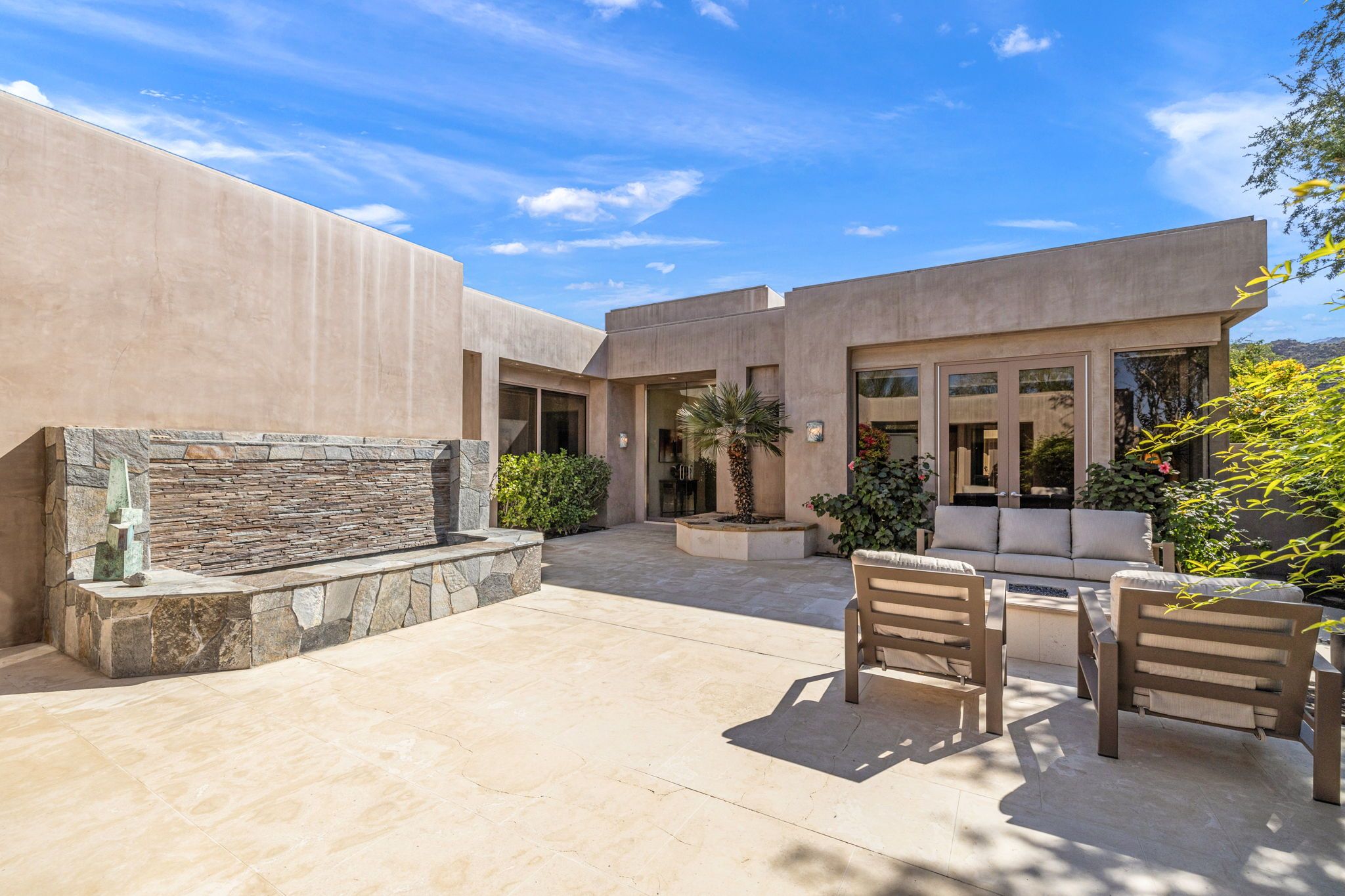 Un patio extérieur moderne avec un mur en pierre, un espace salon avec une table et des chaises, et un bâtiment doté de grandes fenêtres.