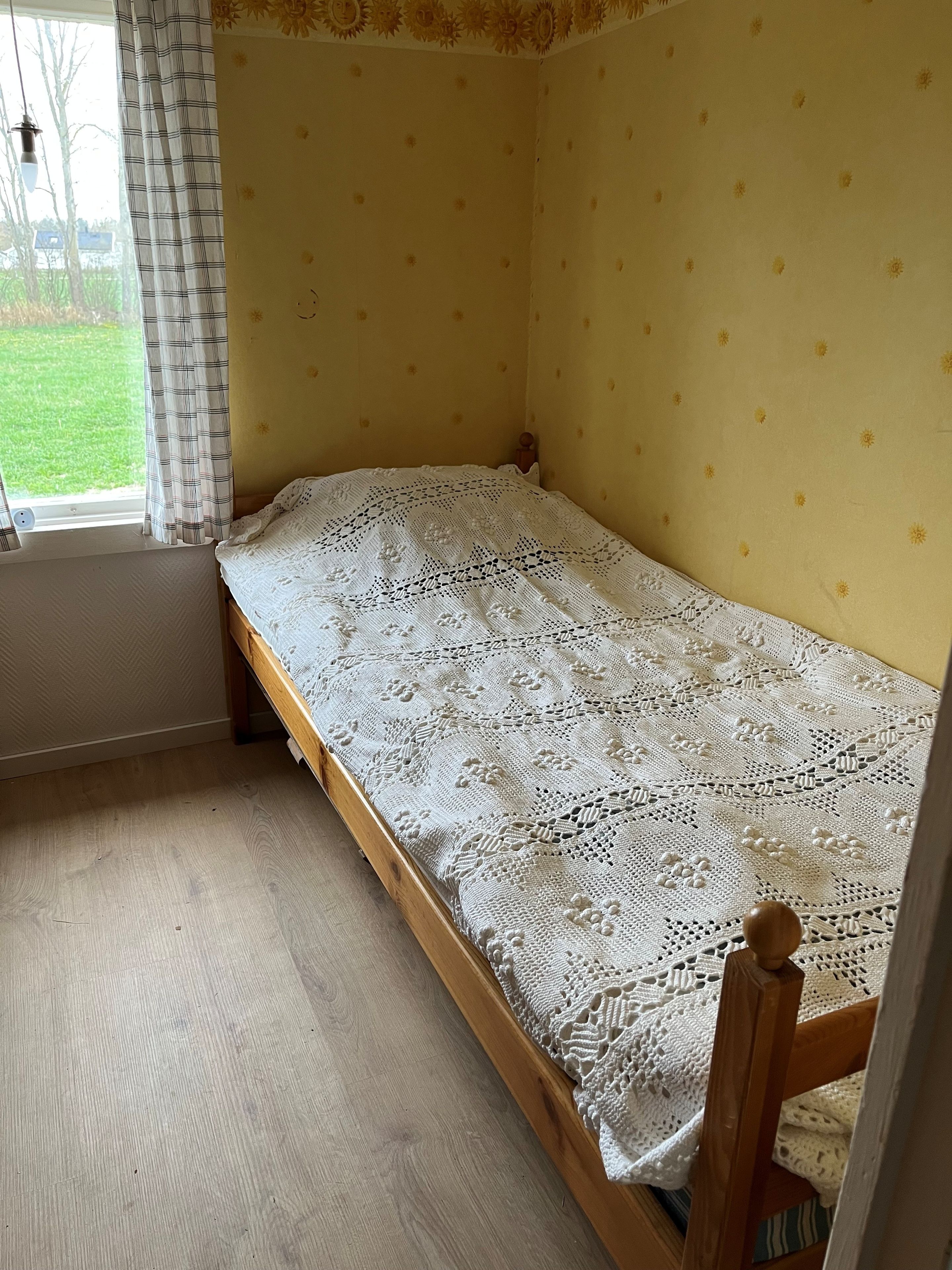 Een eenpersoonsbed met een witte kantbedekking in een kamer met gele wanden en een raam met gordijnen.