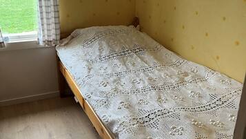 Een eenpersoonsbed met een witte kantbedekking in een kamer met gele wanden en een raam met gordijnen.
