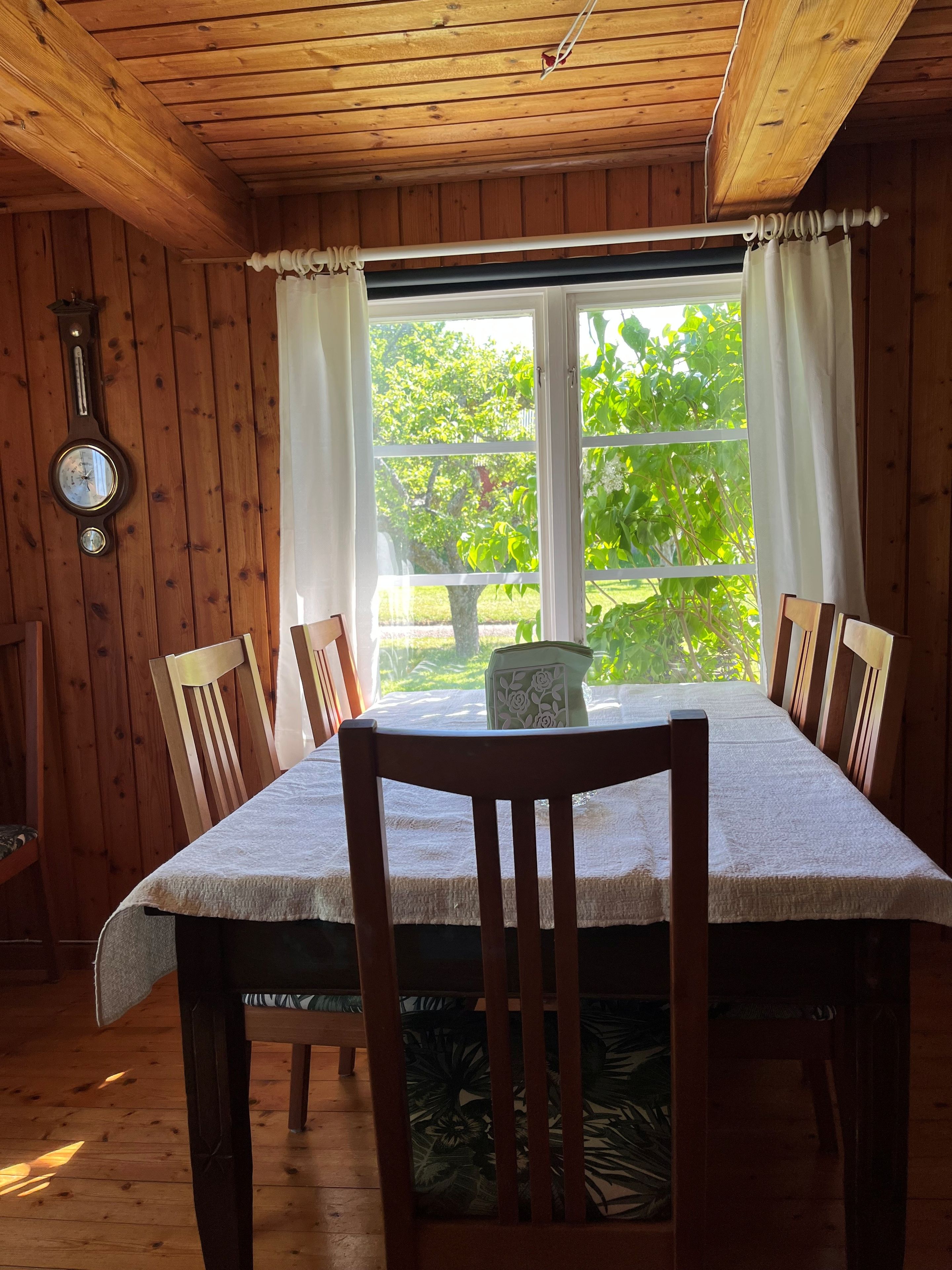 A wooden interior with a dining table, chairs, a clock, and a window with curtains.