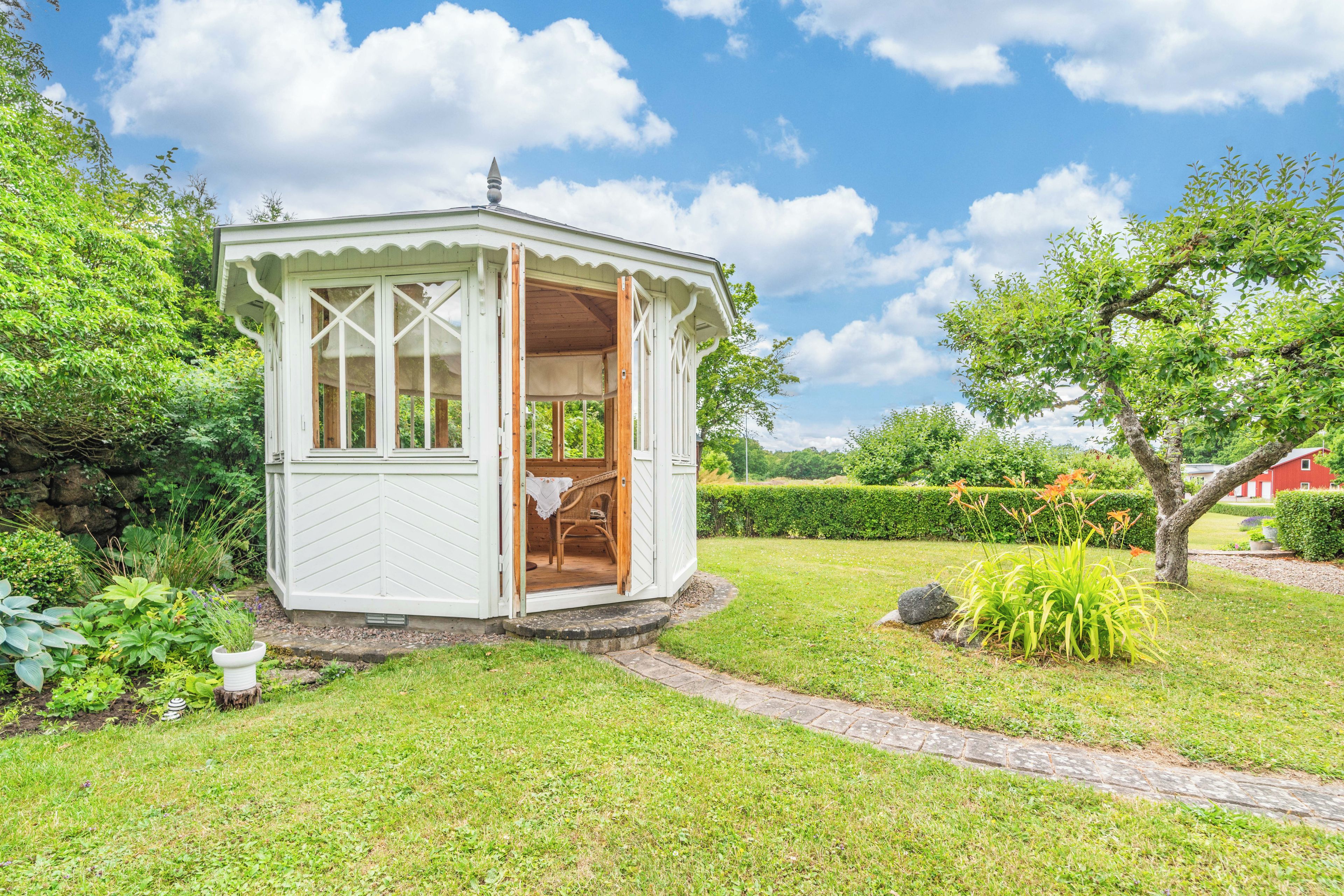 A small, white, open-sided structure with a wooden interior, surrounded by lush greenery and a clear sky.