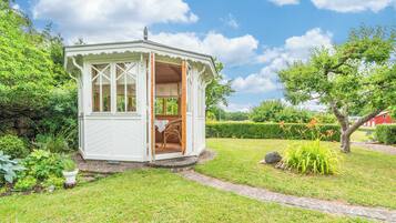 Een klein, wit, open gebouw met een houten binnenkant, omringd door weelderig groen en een heldere lucht.