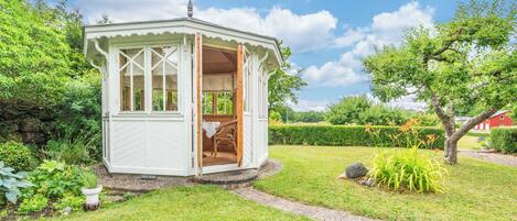 A small, white, open-sided structure with a wooden interior, surrounded by lush greenery and a clear sky.