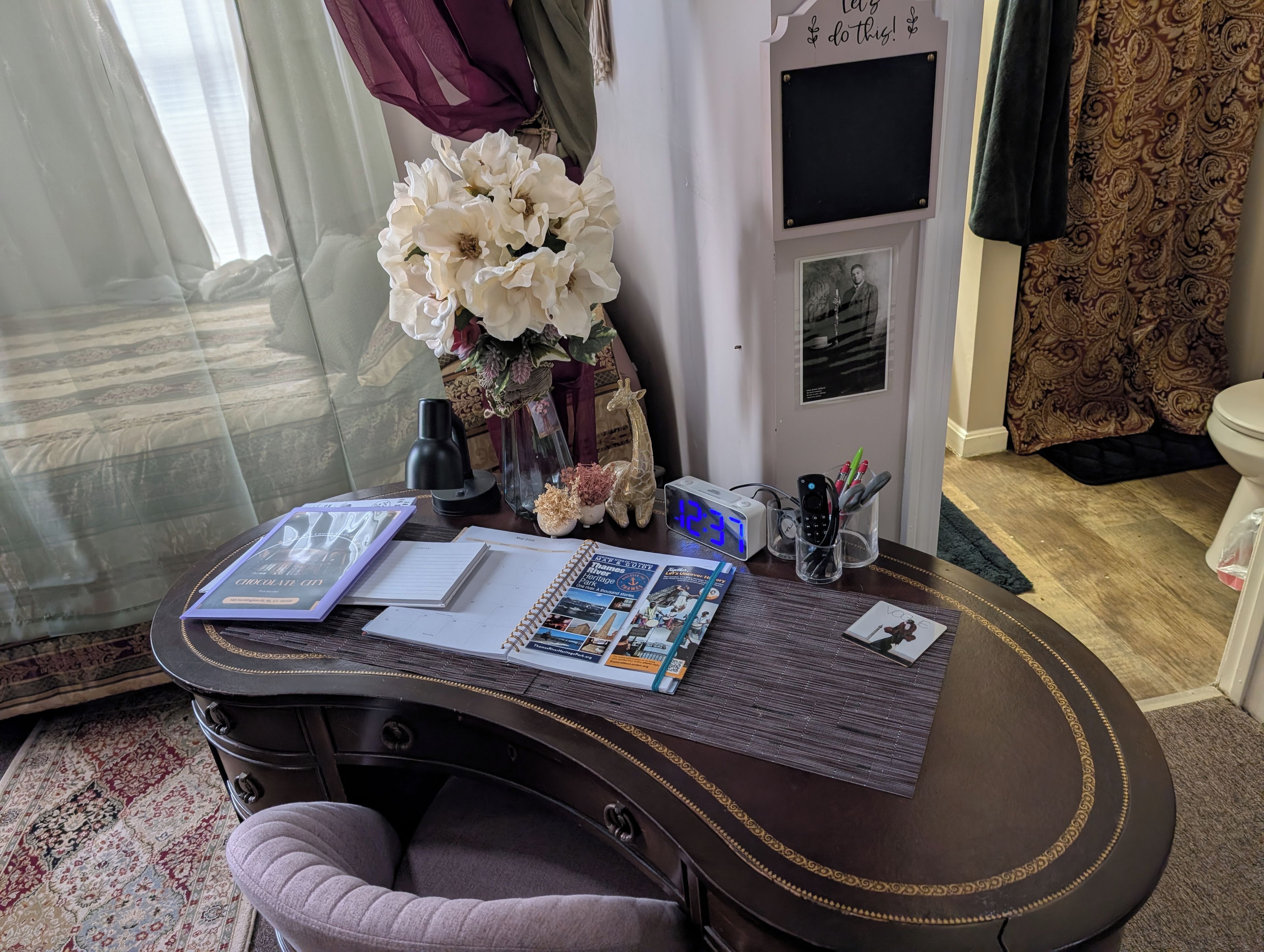 Una habitación con un escritorio, una silla, un reloj, un jarrón con flores y un cuadro en la pared.