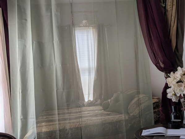 Et værelse med seng, et vindue med gardiner, et natbord med lampe og en vase med blomster.