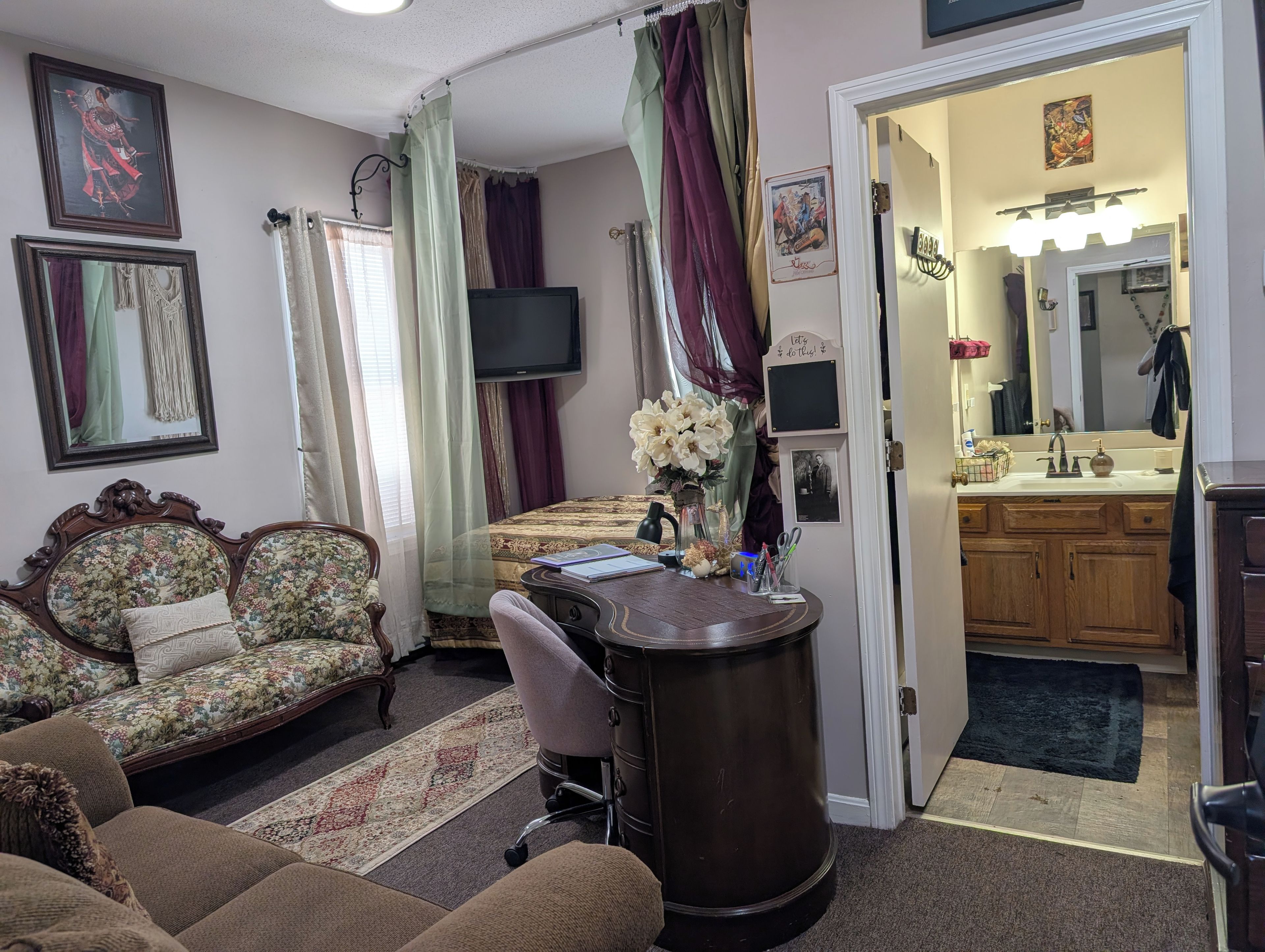 A room with a bed, a desk, a chair, a television, and a bathroom visible through an open door.