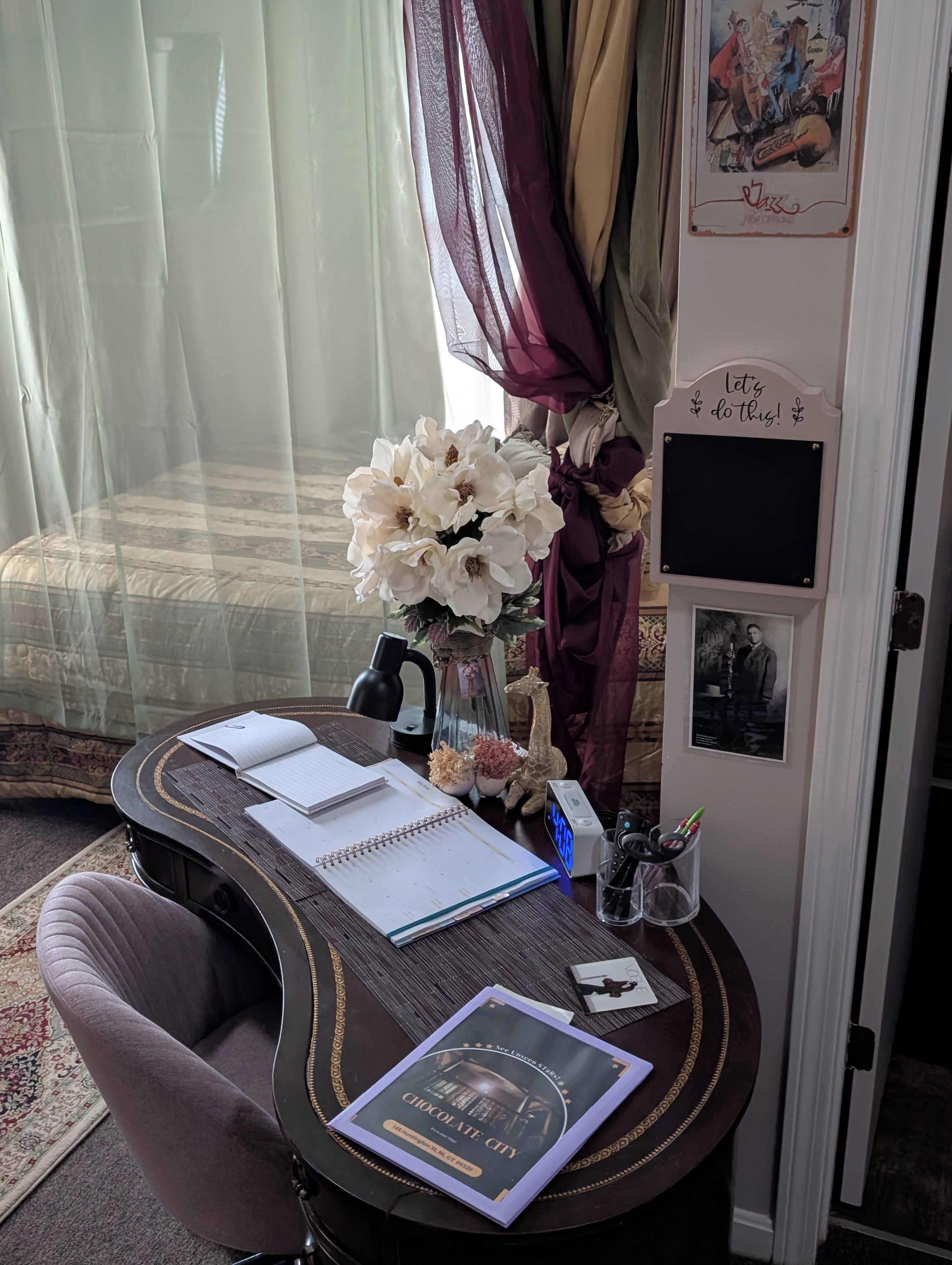 Ein Zimmer mit einem Bett, einem Schreibtisch mit Notizbuch, einem Buch, einer dekorativen Vase, einer Uhr und einem Wandbild sowie einem Schild.